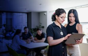 Two women holding an Ipad and talking on a background of three people sitting by a table