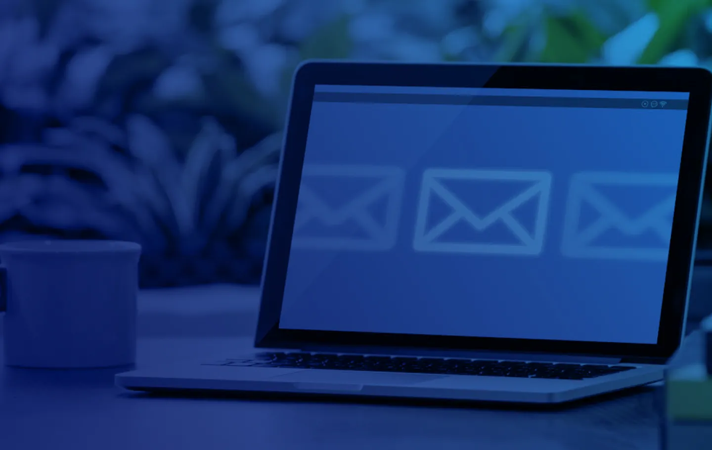 A laptop on a table with three email images on the screen, next to it a white cup