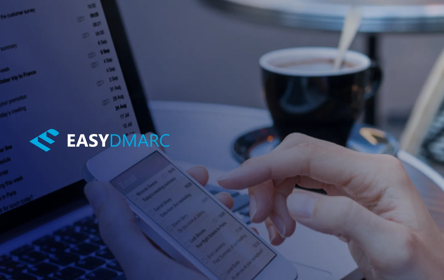 A laptop on a desk, cup on it's right side and mobile  phone with EasyDMARC logo on the picture